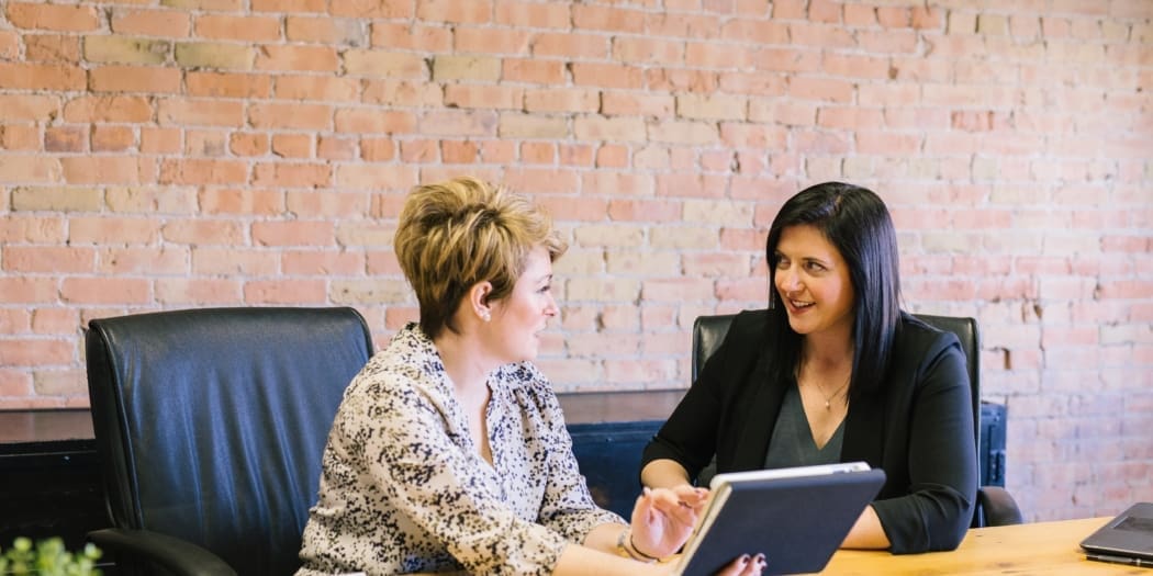 Two business woman smile discuss accounting at a desk