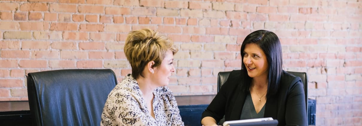 Two business woman smile discuss accounting at a desk