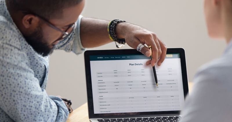 Black male consultant points to a laptop screen with bookkeeping plan details. He is discussing options with a potential customer, who is out of frame.