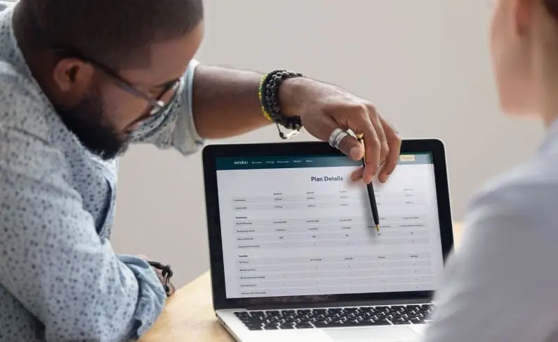 Black male consultant points to a laptop screen with bookkeeping plan details. He is discussing options with a potential customer, who is out of frame.