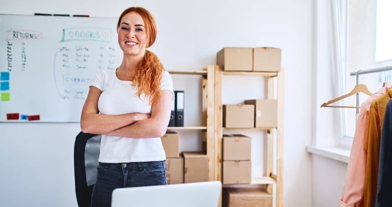 A white, red-head female business owner stands proudly in her office, surrounded by packages