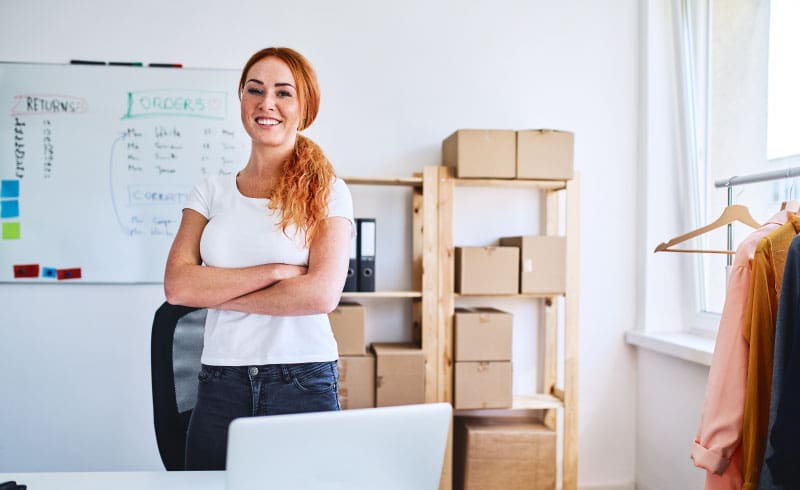 A white, red-head female business owner stands proudly in her office, surrounded by packages