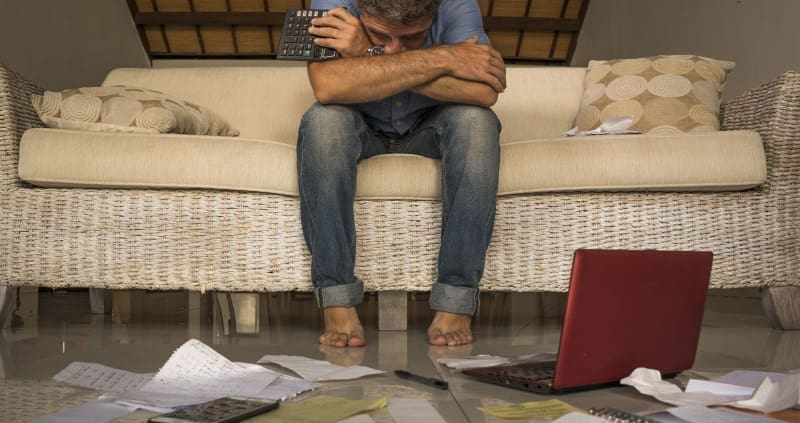 White male business owner, hunched over on his couch, staring at a pile of documents with a calculator in hand