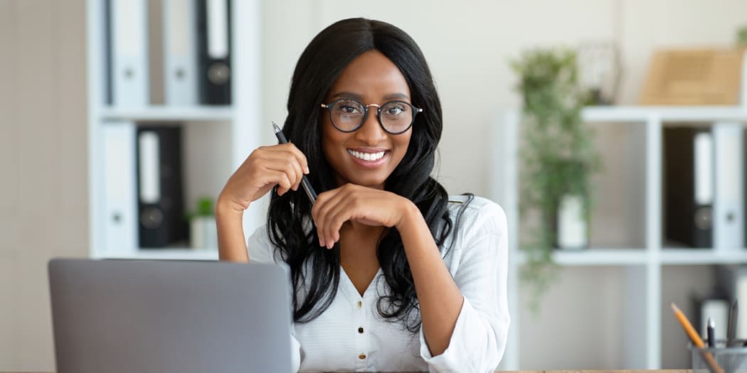 Portrait of cheerful black business lady sitting at her desk with laptop, smiling at camera in office. running her small business
