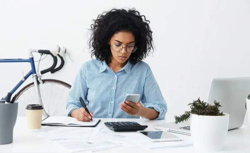 a woman looking at a phone while writing in a notebook